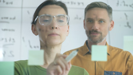 Professional woman with glasses marking glass board, arranging sticky notes while male colleague...