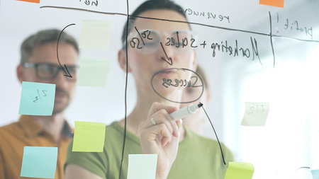 Professional team brainstorming, sharing strategy on glass whiteboard with colorful sticky notes, working collaboratively in bright office spaceの写真素材