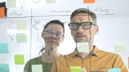 Business colleagues brainstorming, writing strategy notes on glass board, collaborating with...