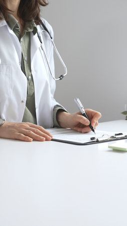Professional female doctor wearing white lab coat and stethoscope documenting patient information on clipboard near laptop in clinic workspace. Medicine and health careの写真素材