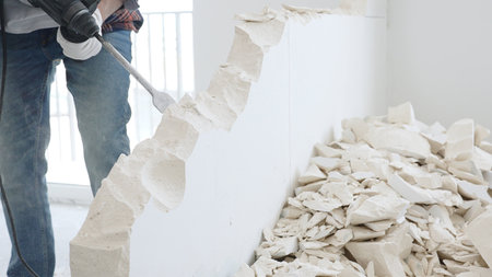 Close up of unknown male construction worker wearing red checkered shirt jeans and protective gloves, is demolishing white wall with rotary hammer drill, generating debris on the floorの写真素材