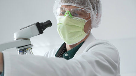 Scientific professional wearing green protective equipment examining green filled test tube near the microscope, performing laboratory research with precision. Medicine, health care and science conceptの写真素材