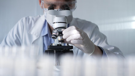 Man scientist researcher wearing white gloves, protective glasses and medical mask is using microscope in modern laboratory, portrait view. Low key lighting photo. Science and Medicineの写真素材