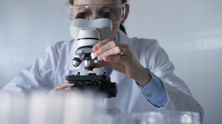Woman scientist wearing lab coat and protective glasses is using microscope in modern laboratory setup. Low key lighting photo. Science and Medicineの写真素材