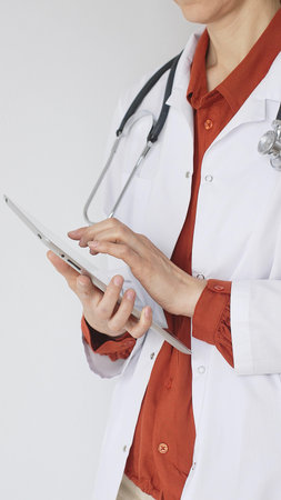 Close up of professional woman doctor, wearing white coat over a brick-red blouse and a stethoscope, is using a digital tablet, suggesting innovative healthcare practices in medicineの写真素材