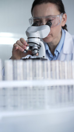 Woman scientist wearing lab coat and protective glasses is using microscope in modern laboratory setup. Low key lighting photo. Science and Medicine conceptsの写真素材