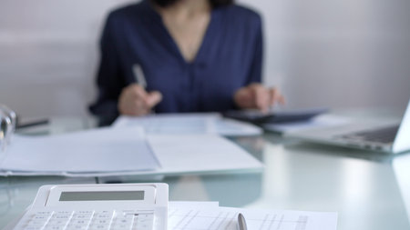 White calculator, pen and documents are lying on accountants desk. Businesswoman wearing blue dress is working with documents on the background. Business and audit conceptの写真素材