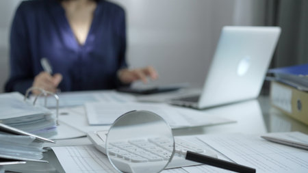 Magnifying glass, white calculator, pen and documents are lying on accountants desk. Businesswoman wearing blue dress is working with documents on the background. Business and audit conceptの写真素材