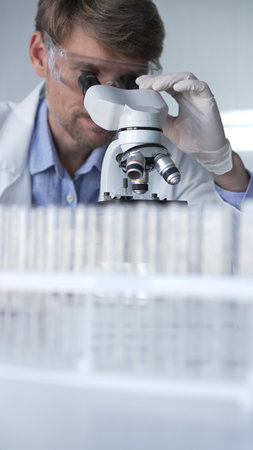 Man scientist wearing lab coat, white gloves and protective glasses using microscope doing research in modern laboratory, portrait view. Low key lighting photo. Science and Medicineの写真素材