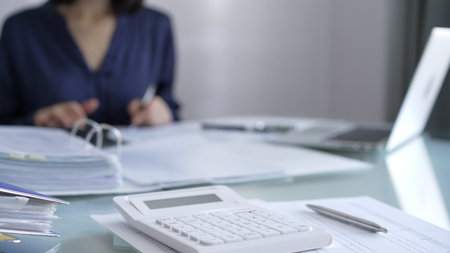 White calculator, pen and documents are lying on accountants desk. Businesswoman wearing blue dress is working with documents on the background. Business and audit conceptの写真素材