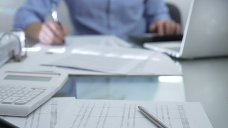 Calculator, pen and documents are lying on accountants desk. Businessman wearing blue shirt is working with documents on the background. Business people, audit conceptの写真素材