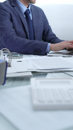 Businessman wearing blue suit is working with laptop, calculator and documents at the glass table in office set. Low lighting, close-up vertical view. Business people and auditの写真素材