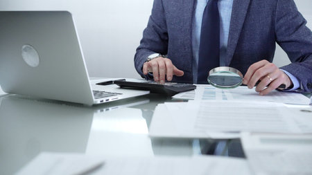 Businessman wearing blue suit is analyzing financial reports with magnifying glass and using...