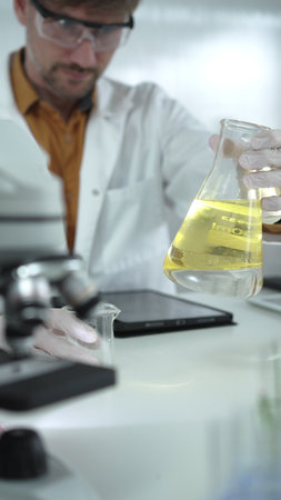 Man scientist wearing white protective gloves, and glasses is holding a yellow chemical solution inside an Erlenmeyer flask in a laboratory, vertical portrait. Science and medicineの写真素材
