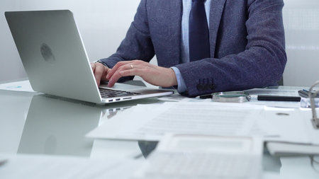 Businessman wearing blue suit is working with laptop, calculator and documents at the glass table in office set. Low lighting, close-up view. Business people and audit conceptの写真素材