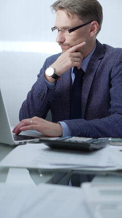 Businessman wearing blue suit is thinking while working with laptop, calculator and documents at the glass table in office set. Low lighting, vertical portrait. Business people and audit conceptの写真素材
