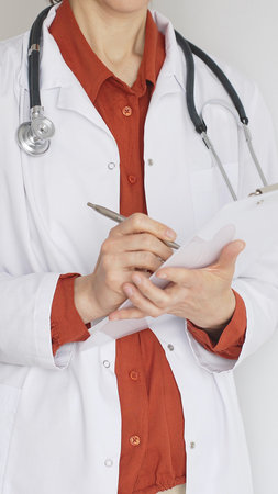 Close up of female professional doctor wearing stethoscope and white coat over brick red blouse is standing and writing on a clipboard, representing medical expertise and care. Medicine conceptの写真素材