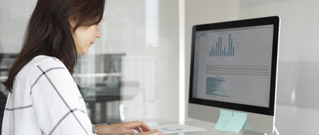 Businesswoman at glass office desk analyzing charts and graphs on computer screen, reviewing financial reports and performance data for planning. Business people conceptの写真素材