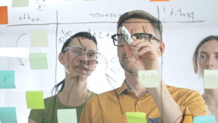 Business colleagues brainstorming, writing strategy notes on glass board, collaborating with...