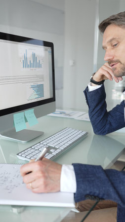Businessman in a blue jacket working on computer, typing and writing notes in modern office, managing tasks, multitasking. Business people conceptの写真素材
