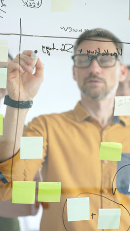 Business team collaborating on a project, using a glass board with sticky notes and marker to brainstorm ideas and develop a strategic plan in office spaceの写真素材