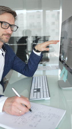 Businessman in a blue jacket analyzing financial data on computer, working in modern office,...