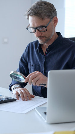 Businessman working in office using calculator and magnifying glass analyzing financial documents, performing accounting and financial analysis. Audit and taxes, business conceptの写真素材