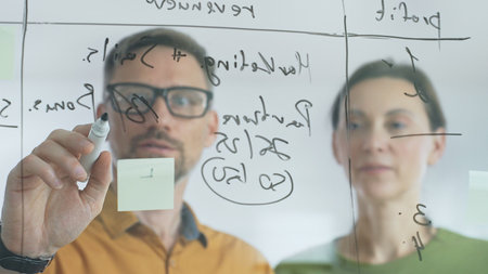 Business people brainstorming near glass board, using sticky notes for strategic planning, writing...