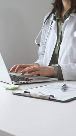 Doctor woman wearing white lab coat over green shirt, and stethoscope, is typing on laptop, documenting patient information with clinical precision at workstation. Medicine and health care conceptの写真素材