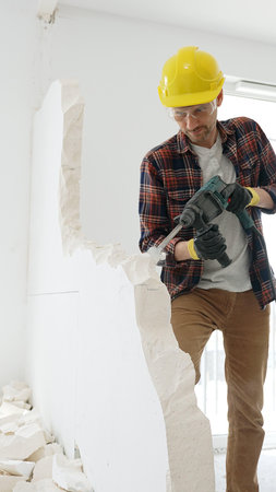 Male construction worker wearing red checkered shirt, yellow protective helmet, is demolishing white wall with rotary hammer drill, generating dust, medium long shot. Renovation projectの写真素材