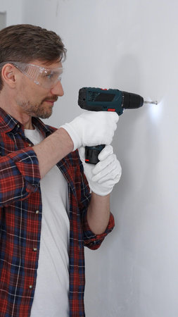 Middle aged male professional worker wearing red checked shirt and white gloves, is drilling hole in...