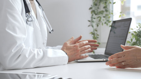 Doctor in lab coat and stethoscope gesturing while explaining diagnosis to patient using laptop in medical office. Medicine and health care conceptの写真素材