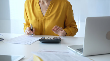 Female accountant wearing yellow shirt is sitting at white desk and using calculator while taking...