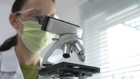 Female scientist wearing green mask and protective glasses using microscope in laboratory, doing...