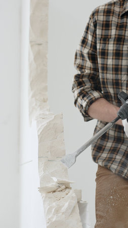 Close up of unrecognizable male construction worker wearing beige checkered shirt and protective gloves, is demolishing white wall with rotary hammer drill, generating dust. Renovation conceptの写真素材