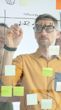 Business team collaborating on a project, using a glass board with sticky notes and marker to...