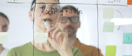 Professional businesswoman wearing glasses writing on glass board, marking strategic plan with...