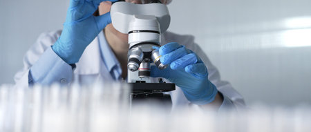 Woman scientist wearing lab coat, white protective gloves and glasses, is using microscope in modern...