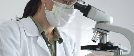 Female scientist examining microscope slide, wearing protective gear while conducting medical...