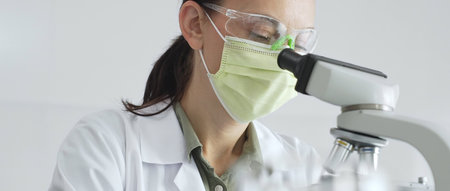 Female scientist examining specimen, placing slide under microscope while wearing green protective...