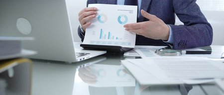 Businessman wearing blue suit is working with laptop while examining financial data, using charts...