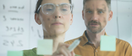 Professional woman with glasses marking glass board, arranging sticky notes while male colleague...