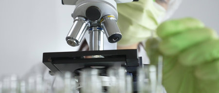 Scientist wearing green gloves and a face mask selecting a test tube with transparent liquid from a...