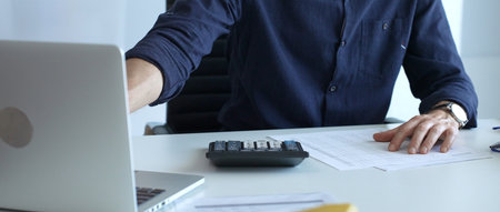Businessman in a blue shirt examining financial documents at an office desk, using a calculator...