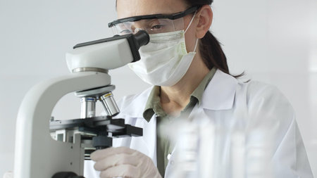 Female scientist examining microscope slide, wearing protective gear while conducting medical...