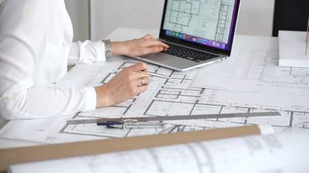 Architect designing residential floor plans, reviewing blueprints on a laptop and using a compass to...