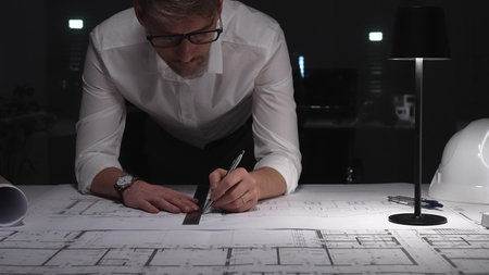 Architect working on blueprints late at night, focusing on construction plans and project...