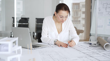 Professional female architect working on building plans in a modern office, drawing blueprints with...