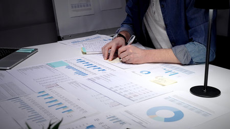 Man actively working on a busy desk, reviewing financial reports and data analysis on paper...