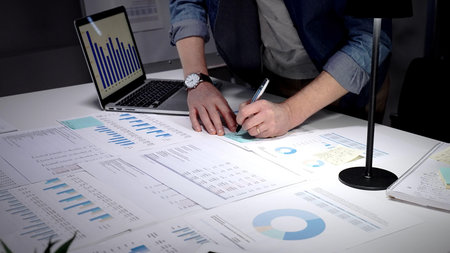 Person analyzing financial data, reviewing business reports and charts on a desk during a late work...